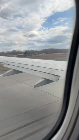 The video captures a view from an airplane window during takeoff. The main subject is the airplane's left wing, clearly visible from the passenger cabin. The wing features several flaps and other aerodynamic surfaces.

The plane is on a concrete runway, which quickly recedes beneath the wing as the aircraft accelerates. Beyond the runway, there's a grassy field transitioning into a distant line of trees. Some industrial-like buildings are visible in the far background.

The sky is partly cloudy, with patches of blue sky visible through the clouds. The lighting suggests it is daytime. The sound of jet engines accelerating is prominent throughout the video, indicating the powerful thrust needed for takeoff. The airplane speeds up along the runway before lifting off the ground towards the end of the clip. No people or visible text are discernible in the frame. The location is Bolshoye Savino, Russia.