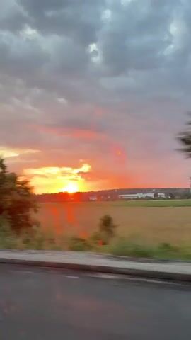 The video captures a moving view of a sunset behind trees and fields. The sky is filled with dramatic clouds, ranging from dark gray to vibrant orange and pink hues. The sun, a bright orb, is visible low on the horizon, casting a warm glow over the landscape. In the foreground, trees and foliage are blurred due to motion, suggesting the footage was filmed from a vehicle. The ground in the immediate foreground appears to be a road or path, with some asphalt visible.