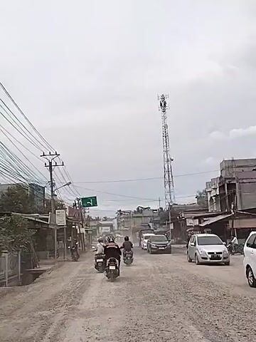 A dusty, unpaved road with motorcycles and cars traveling on it is shown. A tall cell tower is visible in the background, along with buildings and power lines. The sky is overcast.
