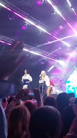 A concert is taking place with a stage bathed in purple lighting. Two women are performing on stage. One woman, with blonde hair, is wearing a white top and a red skirt, and is singing into a microphone. The other woman, with dark hair, is wearing a white top and a blue skirt, and is also singing. They are both dancing. A crowd of people is visible in the foreground, holding up their phones to record the performance. The stage is equipped with lighting rigs and a drum set.