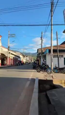 The camera pans up the main street of a small town on a sunny day, moving away from the sidewalk into the street.  Buildings flank both sides of the street. There are cars parked and moving on the street and people walking on the sidewalks.  A man on a motorcycle turns to look at the camera.  Power lines crisscross the blue sky above.  Green, hilly terrain is visible in the distance at the end of the street.
