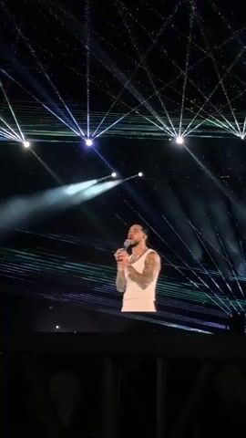 A man wearing a white tank top and white pants stands on a raised platform. He holds a microphone. He raises his arms. He is singing. There are chairs, a fan, and a small table on the platform. There are white lights and laser beams above him. The background is dark.