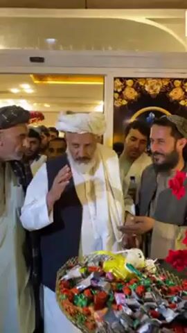 The video captures an indoor scene in Kandahar, Afghanistan, featuring a group of men, primarily dressed in traditional Afghan attire. The setting appears to be an entrance or doorway, well-lit by artificial overhead lighting, suggesting it is evening or night.

The central figure is an older man with a long white beard, wearing a white turban, a white traditional tunic, and a dark waistcoat. He is actively gesturing with his hands, appearing to speak or direct the group as he moves forward.

Surrounding him are several other men, also in traditional clothing, including various head coverings and tunics. One man to the central figure's right (viewer's left) is wearing a dark turban and a light-colored tunic with a dark shawl. Another man to the central figure's left (viewer's right) is holding a red rose.

In the foreground, a large, dark basket is prominently displayed, filled to the brim with an assortment of individually wrapped, brightly colored sweets or candies, along with several red roses. This arrangement strongly implies a welcoming ceremony or a celebratory event.

The background includes glass door panels with decorative gold patterns on a dark surface, and some indistinct text is visible as part of the decor. The men seem to be entering the space, with the central figure leading or addressing the others. The overall atmosphere suggests a formal gathering or a significant arrival.