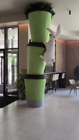 A white rabbit figurine is climbing a stack of three oversized, bright green cups with black lids. The cups appear to be decorative or part of a large installation. The rabbit is positioned on the middle cup, reaching upwards towards the top cup. The scene is indoors, with a tiled floor and large glass doors visible in the background. There are also tables and chairs in the background, suggesting a cafe or restaurant setting.