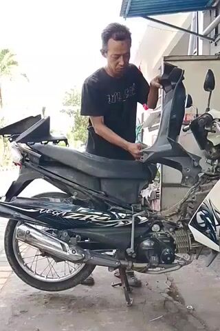 A man in a black t-shirt is standing next to a disassembled white and black motorcycle. The man is holding a black plastic motorcycle fairing in his hands. He appears to be in the process of either removing or installing the fairing onto the motorcycle. The motorcycle is on its kickstand. The environment appears to be outdoors, possibly a workshop or garage, with a blue roof overhead and some foliage visible in the background.