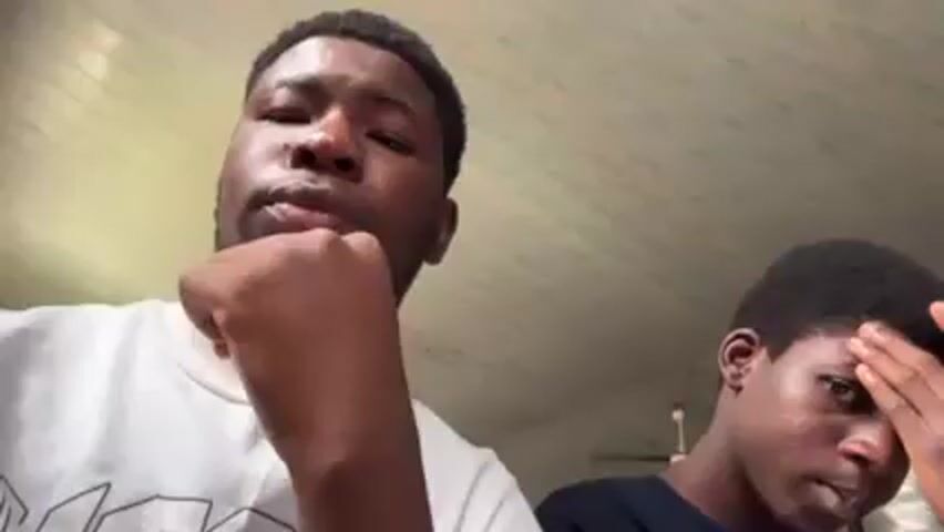 Two young men are looking directly at the camera. The man in the foreground is wearing a white t-shirt and has a close-cut haircut and a short beard. He briefly rests his hand under his chin, then puts it down. The man in the background is wearing a dark t-shirt and is touching his forehead. The background is a white ceiling with thin brown stripes.
