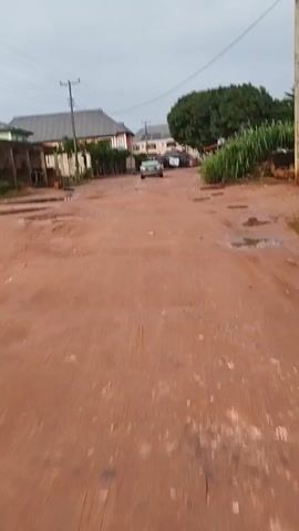 A car drives down a dirt road. The road is wet and muddy. There are puddles of water on the road. There are houses and trees on the side of the road. The sky is cloudy.