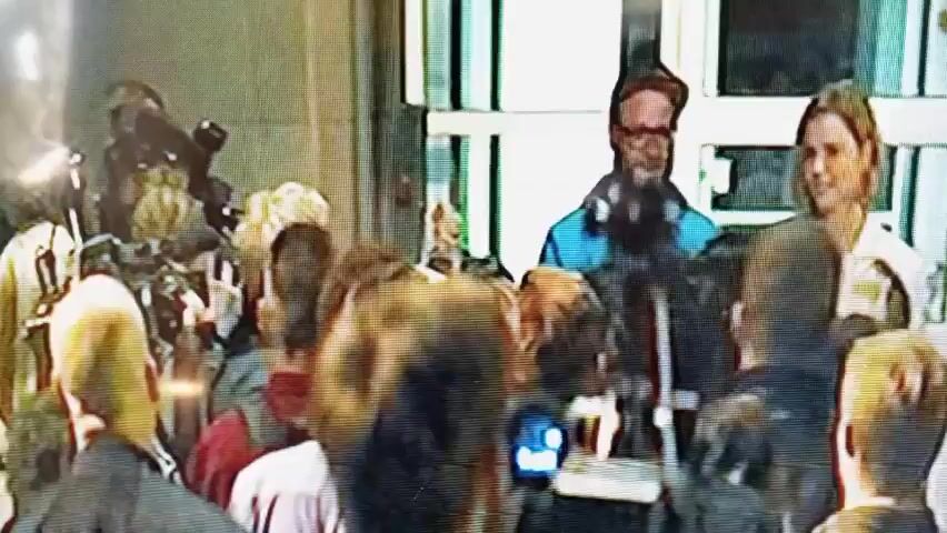 The video begins with a close-up of a diverse group of people, their faces blurry and illuminated by an unseen light source. The camera then shifts focus to a man with a beard and glasses, who appears to be speaking to a woman with shoulder-length blonde hair. The woman smiles as the man gestures towards her. The video briefly shows another man wearing glasses and a blue shirt. The overall environment appears to be indoors with a yellowish tint, suggesting artificial lighting or a specific color filter. The footage is grainy and of low resolution.
