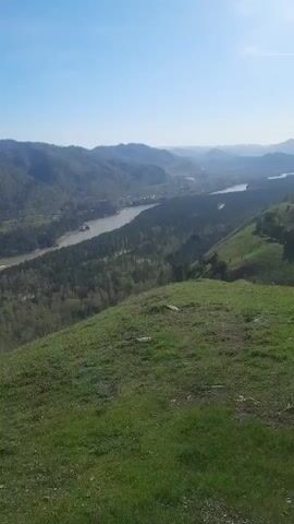 This video captures a panoramic view of a scenic river valley in Aya, Russia, on a clear, sunny day. The camera pans across a vast landscape, starting with a wide shot of a winding river flowing through a forested valley, flanked by tree-covered mountains in the distance. The vibrant green of newly grown grass covers the foreground slope.

As the camera continues to pan, it reveals a distinct rock formation or cliff face where several people are gathered, enjoying the view. Beyond this, a small town or village is visible nestled in the valley alongside the river. The final shot frames a simple wooden bench situated on a dirt patch, offering a perfect vantage point to overlook the expansive valley and the distant mountains. The bright blue sky is mostly cloudless, and the sunlight casts clear shadows from the bench, indicating good weather. No text is visible in the video.