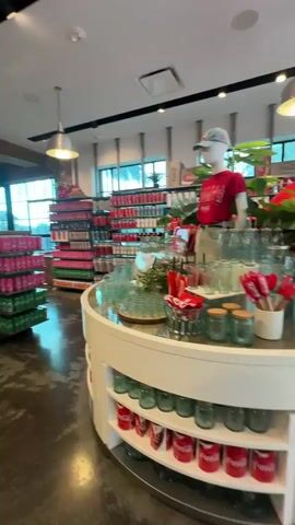 The video shows the interior of a Coca-Cola retail store. The camera pans from a display of Coca-Cola products to show shelves of Coca-Cola themed merchandise, including bottles, bags, and clothing. People are seen walking through the store. The environment is well-lit with modern retail design elements.