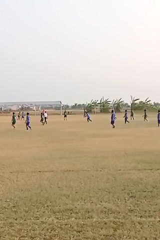 A wide shot captures a group of young people playing soccer on a dry, grassy field. The sky is overcast and pale, suggesting an outdoor daytime setting. The players, dressed in various colored jerseys, are spread across the field, some in motion, others stationary. Two goals are visible at opposite ends of the field, indicating the boundaries of the game. In the background, a long, low building with a corrugated roof and some trees are visible. The overall impression is of a casual, informal soccer match taking place in a somewhat arid environment.