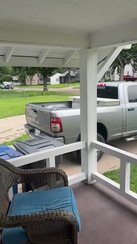 This video showcases a residential street scene viewed from a porch. The main subjects are a silver Ram pickup truck parked on a paved driveway, and a portion of the house's porch in the foreground. The porch features white painted railings and supporting posts, with a wicker chair and blue cushion visible on the left. Beyond the truck and driveway, the scene opens up to reveal a suburban neighborhood. Several houses, mostly of a traditional architectural style, are visible, set back from the street with well-maintained lawns and mature trees. A sidewalk runs along the front of the houses, leading away from the viewer.

The activity appears to be static, with the truck parked and no people or significant movement captured in the frame. The time of day seems to be daytime, with bright natural light casting shadows, suggesting a clear or partly cloudy day. The environment is green and lush, indicating a temperate climate, likely spring or summer. The setting is a quiet residential street in Des Moines, Iowa, United States, characterized by its orderly homes and landscaping. No explicit emotions are conveyed, but the overall impression is one of peaceful, everyday suburban life. There is a license plate visible on the back of the truck, but the text is not legible. There is also a toolbox visible in the bed of the truck.