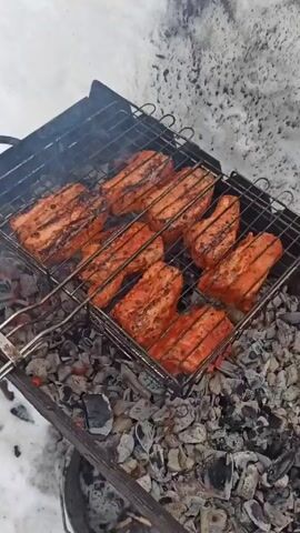 The video shows a close-up of pieces of meat grilling on a barbecue grill over hot coals. The meat is arranged in rows on the grill. The grill is placed on a metal charcoal container. There is snow on the ground around the grill. The meat appears to be seasoned and is beginning to char on the grill. Smoke is rising from the grill.