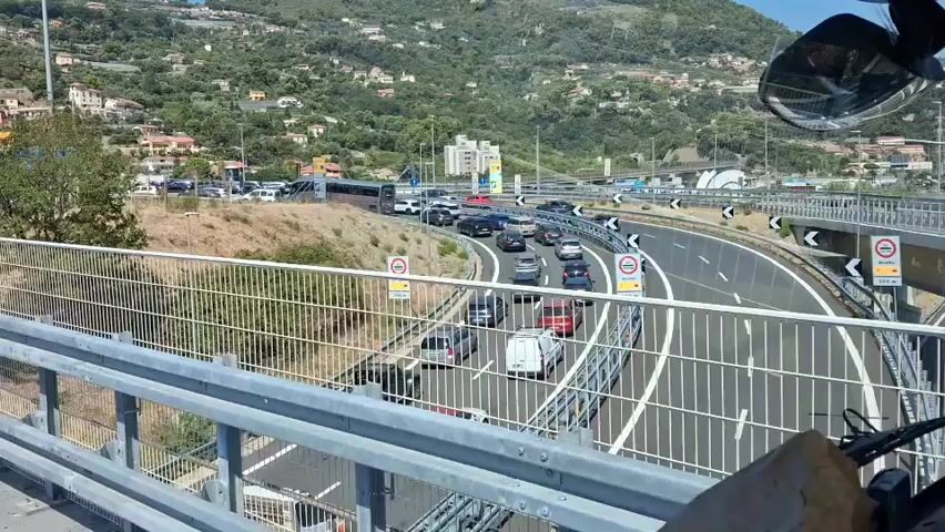 A drone shot captures a highway with moderate traffic. In the foreground, a grey Tesla is visible. The camera pans to the right, showing a highway with several lanes of cars and trucks. A large electronic sign displays "40" with a circle around it, indicating a speed limit. In the background, a hillside is dotted with houses and trees. The highway leads to a toll plaza, where a long line of vehicles is forming. The sky is clear and blue.