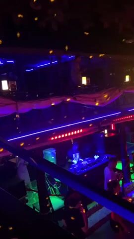 The video shows a DJ performing in a nightclub. The DJ is mixing music on a console, illuminated by blue and red lights. People are seen standing and watching the DJ. The venue is decorated with disco lights, and there are multiple levels. There are blue, red and white lights lining the bar and the ceiling. A red cloth is draped on the ceiling.