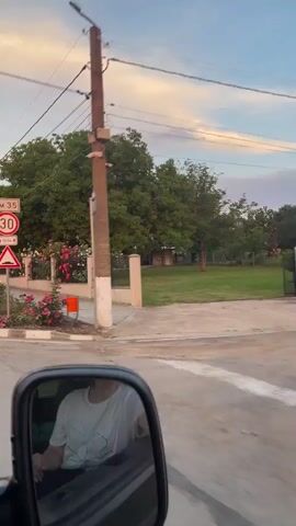 The video is filmed from inside a moving vehicle, looking out the passenger window and reflecting the driver in the side mirror. The driver, a person with short brown hair, wearing a white t-shirt, is visible. The vehicle is on a street lined with houses and rose bushes. There is a street sign indicating "KM 35" and a speed limit of "30," along with a speed bump warning. The houses are two stories high, with reddish-orange roofs, and are surrounded by lush rose bushes in various shades of pink and red. The sky is a blend of pale blue and orange, suggesting either sunrise or sunset. A light pole is also visible on the right side of the frame.