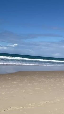 The video shows a wide shot of a sandy beach on a sunny day. The ocean is visible in the background with waves breaking on the shore. The sky is blue with scattered clouds. Several people are walking on the beach. In the first few seconds, a man in dark shorts walks towards the water. Then, a person wearing a red hat, a light-colored jacket, and dark shorts walks away from the camera, carrying a bag. Further down the beach, a woman with blonde hair, wearing a white top and shorts, walks towards a group of modern high-rise buildings that line the coast. There are other people and a red flag visible in the distance along the beach. The camera pans to the right, revealing more of the buildings and the beachgoers. The video ends with a view of the beach and the cityscape.