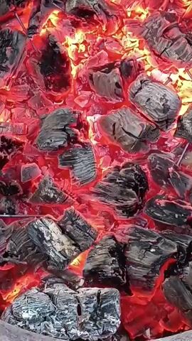 A close-up shot shows glowing red-hot charcoal briquettes. The briquettes are charred and black on the outside, with bright red embers visible through cracks and gaps. The heat from the coals creates an intense red glow that illuminates the scene. The camera is positioned directly above the coals, providing a top-down view. The overall impression is one of intense heat and the process of combustion.