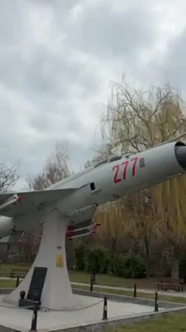 The video shows a gray MiG-21 fighter jet mounted on a pedestal in a park. The jet has a red "277" painted on its side and is equipped with missiles under its wings. The pedestal is square and made of a light-colored stone. There is a plaque on the front of the pedestal. The surrounding park has a green lawn, trees, and a cloudy sky overhead.  There are no people visible in the video.