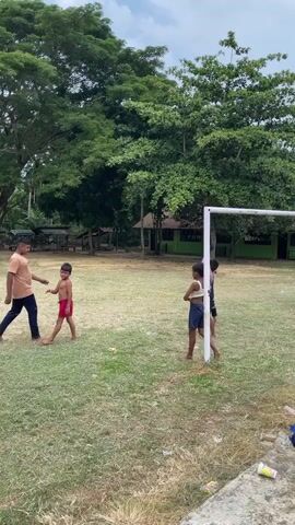 A group of people, mostly children, are playing soccer on a grassy field with trees in the background. The weather appears overcast. One adult man is present on the field with the children. A soccer goal is visible in the background. The children are wearing casual clothing, and most are barefoot. One child is kicking the soccer ball.