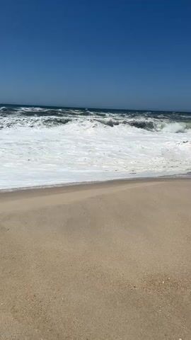 This is a short video clip of a beach in Torreira, Portugal. The scene features a sandy beach in the foreground, with the ocean and a clear blue sky dominating the background. The waves are breaking with white foam, indicating a moderate surf. The sun is shining brightly, suggesting it is daytime, likely late morning or early afternoon given the angle of the light. There are no people or objects visible in the frame. The overall mood is tranquil and natural, capturing the essence of a sunny day at the coast. No text is visible in the video.