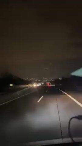 The video shows a car traveling on a highway at night. The road is wet, and there are other cars visible in front. In the distance, the lights of a city can be seen. The sky is dark and cloudy. A small electronic screen is visible on the lower right corner of the frame.