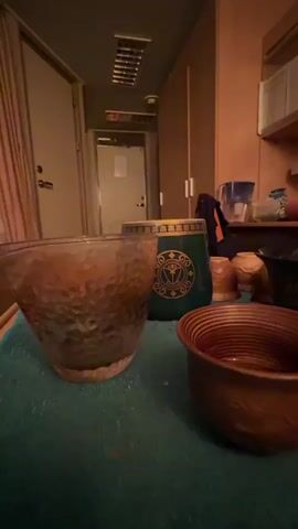 In a dimly lit room, a hand is seen pouring a dark liquid from a small pitcher into a hammered copper bowl. The bowl is filled about halfway. To the left of the bowl is a larger, wider hammered copper bowl, also partially filled with the same dark liquid. Behind these bowls is a tall, cylindrical object with a green exterior and an ornate gold emblem featuring a crest with a shield and a laurel wreath. Several smaller, light-brown ceramic cups are arranged behind the green cylinder. The scene appears to be set in a room with wooden cabinetry and a closed door in the background. The lighting suggests it could be evening or a room with limited natural light. There is no visible text in the frame. The activity appears to be the preparation of a beverage, possibly tea or coffee, in a rustic setting.