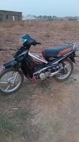 The video presents a stationary, black motorcycle, possibly a scooter or underbone type, parked on a dirt surface. The background consists of dry grass and sparse vegetation with a few unfinished or abandoned building structures visible in the distance. The motorcycle has silver-colored wheels, a dark seat, and pink accents on its lower body. The sky appears overcast or the video may have been taken during dusk.