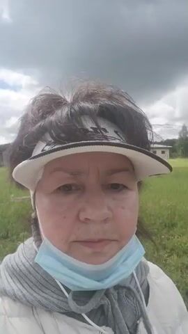 A woman is filmed from the chest up, outside in what appears to be a grassy area with a building in the background. She is wearing a white visor, a light-colored jacket over a gray scarf, and a light blue surgical mask. Her hair is dark and pulled back. The sky is cloudy. She is looking at the camera and smiling. The video is shaky and appears to be a selfie video.