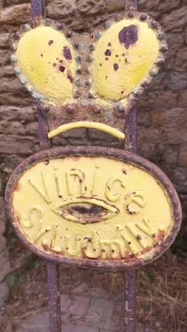 This is a close-up, static video of a rusty metal gate in front of a stone wall. The gate has a decorative element resembling a crowned face with two large, yellow, circular "eyes" that are speckled with brown rust. Below the eyes, there is a curved, thin yellow piece resembling a mouth, which also appears to be rusty. A yellow oval plaque is attached below the mouth. The plaque has embossed lettering that appears to read "Vinice" above what looks like an eye symbol and then "Schlussen". The gate itself is made of vertical rusty bars. The background is a stone wall, and some dry vegetation is visible at the bottom of the frame. There are no people or any discernible actions taking place.