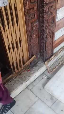 The video shows a close-up view of an ornate wooden doorway with intricate carvings. A metal collapsible gate is pulled shut in front of the doorway, secured with a padlock. There are some scattered flower petals on the floor in front of the doorway, and a metal grate lies on the floor to the right. Above the doorway, there are garlands of flowers and a bell hanging. A glimpse of what appears to be a shrine or altar with draped fabric is visible through the gate. The lighting is somewhat dim, suggesting an indoor or shaded environment.