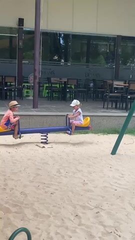 Two young children are playing on a seesaw in a sandpit. The child on the left is wearing a straw hat and colorful shorts, while the child on the right is wearing a white hat and a pink top. They are both holding onto the handles of the seesaw. The seesaw is blue with yellow seats. In the background, there is a building with large windows and tables and chairs. The overall scene is a playground on a sunny day.
