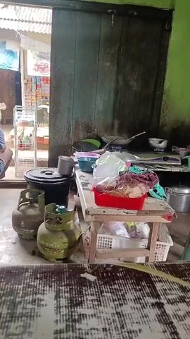 The video shows an outdoor area with two green propane tanks in the foreground. Behind the tanks, there is a wooden table with various items, including pots, bowls, and plastic bags. A person's hand is visible in the bottom right corner. The background features a doorway leading to an area with shelves stocked with items and a green wall. A corrugated metal roof is visible above the doorway. The overall impression is of a cooking or food preparation area.