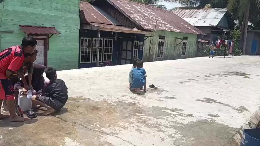 A group of children are in an outdoor concrete area with houses in the background. Four children are gathered on the left side of the frame, three of whom are crouching or sitting and appear to be interacting with an object. One child is standing. Another child is further away in the center of the frame, also crouching. In the far background, another child is visible. The environment appears to be sunny, with some areas of the concrete wet and others dry. Clothing is hanging on a line in the background.