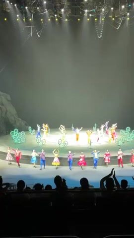 The video shows a stage performance with a large group of performers dressed in colorful costumes, waving and posing for the audience. Some performers are riding bicycles with lit-up wheels. The stage backdrop is a large gray structure resembling rocks. The audience is silhouetted in the foreground, with some people raising their hands. The stage is well lit.