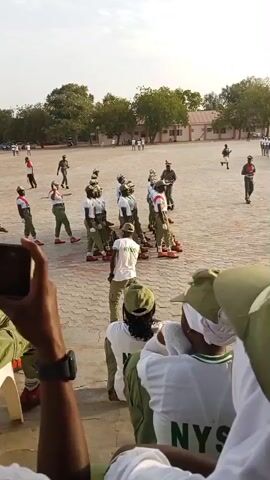 The video shows a group of young people in matching green uniforms and caps participating in a drill or parade on a paved outdoor area. They are arranged in rows, some holding what appear to be sticks or batons. In the foreground, spectators are seated, also wearing green caps and white shirts with green lettering. One person in the foreground is filming the event with a smartphone. The setting appears to be a large open field with buildings and trees in the background. The sky is bright, suggesting it is daytime, possibly late afternoon. The overall mood seems formal and organized, typical of a military or civic service training event. The visible text on the shirts of the people in the foreground reads "NYS," which likely stands for National Youth Service, a program in Nigeria. The location is Ajiwa, Nigeria.