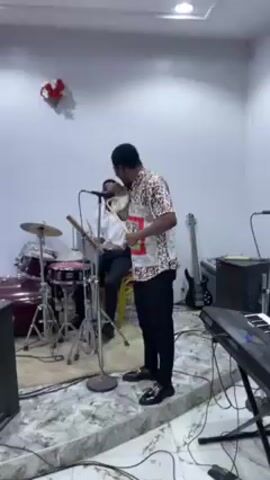 A man sings into a microphone while holding and tapping a tambourine. He is wearing a patterned shirt, black pants, and shiny shoes. Behind him to the left, another man sits at a drum set. The room has musical instruments including a keyboard, bass guitar, and various speakers. The walls are painted white. The floor is covered with a light-colored reflective material, possibly marble or polished concrete. There are decorations on the wall.