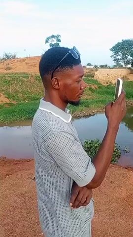 The video shows a man standing near a body of water and using a smartphone. The man is wearing a light gray shirt with short sleeves and has sunglasses on his head. He is holding the phone in his right hand and appears to be looking at the screen. The body of water is a lake or pond with trees and vegetation on the opposite bank. The ground near the man is reddish-brown soil. The sky is partly cloudy.