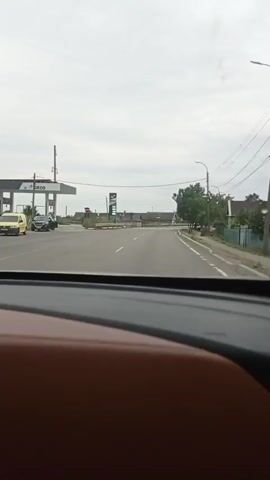 A dashcam view from inside a vehicle shows a road with several cars and a white van. On the left side of the road, there is a gas station with a large sign. There are also houses and trees visible along the right side of the road. The sky is overcast. The vehicle appears to be moving slowly.