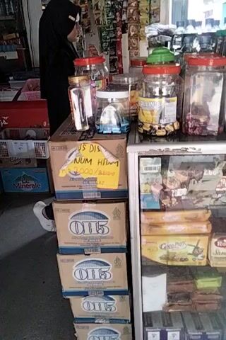 The video shows a cluttered store interior. In the foreground, a person wearing a black head covering and black clothing is partially visible, sitting behind a counter. On the counter are several large plastic containers filled with food items, and a red appliance. Below the counter, several cardboard boxes are stacked, some with blue labels that read "OIL." To the right, a white shoe is visible. In the background, shelves are packed with various packaged goods, including snacks, drinks, and what appear to be canned items. The overall impression is of a small retail establishment with a wide variety of products on display.