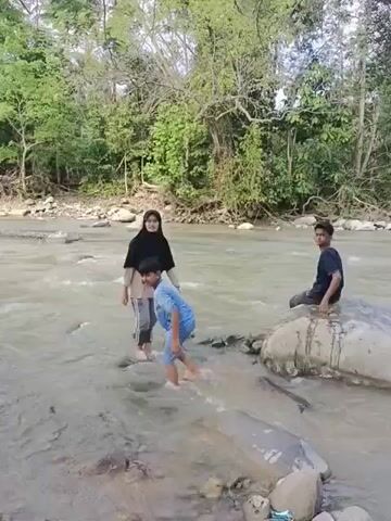 In a natural outdoor setting at Abah Lueng, Indonesia, three young people are enjoying a river. The scene is characterized by a flowing river with clear water and numerous rocks, bordered by dense green trees on both banks, suggesting a lush, forested environment.

A girl wearing a black hijab and light-colored clothing is standing and slowly walking in the river. Near her, a boy in a light blue outfit is actively playing in the water. He is initially on his hands and knees, then attempts a brief handstand or headstand, with his feet pointing upwards, before returning to a kneeling position and splashing. Another boy, dressed in a dark blue t-shirt and dark pants, is seated on a large, smooth rock on the riverbank, observing the others.

The weather appears sunny and bright, indicating daytime. The atmosphere seems relaxed and playful as the individuals engage with the natural surroundings. No visible text is present in the video.