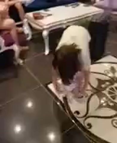 The video shows a young person in a light-colored shirt and shorts bending down to pick up something from the floor. The person's face is obscured by their dark hair. They are on a decorative rug with a circular pattern. In the background, a sofa and a table are visible, suggesting an indoor setting, possibly a living room or a lobby. The person appears to be interacting with an object, but the object is not clearly visible.