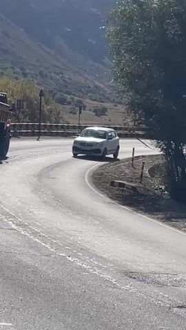 A white hatchback car is driving on a winding asphalt road in a mountainous area. A large orange truck is visible behind the car. The road has yellow and white lane markings. There are trees and rocky slopes on either side of the road, and the sky is partly cloudy. The car appears to be making a turn to the right.