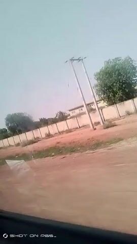 The video shows a view from inside a moving vehicle. The landscape visible outside the window includes a dirt road running parallel to a long, white concrete wall. Several trees are scattered behind the wall. A person wearing a dark shirt or jacket is seen walking on the dirt road at the beginning of the video. The sky is overcast, and telephone poles are visible along the roadside.