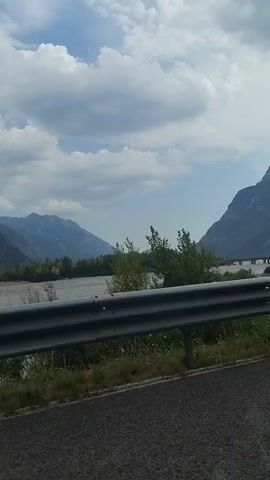 The video shows a view from a vehicle moving along a road, with a guardrail in the foreground and a wide riverbed with a shallow river in the midground. Beyond the river, there are tree-covered slopes leading up to imposing, rugged mountains under a cloudy sky. The overall impression is a scenic, natural landscape, possibly a mountain valley. The audio consists of spoken words, possibly a narration or announcement, which is partially in Italian and mentions "famiglia" (family), "amici" (friends), and "bambini" (children).