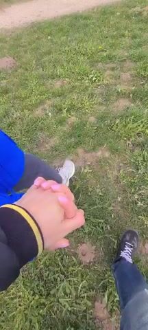 The video shows a first-person perspective of two people holding hands while on the ground outside. The person filming is wearing a black jacket with a yellow stripe on the cuff and grey pants. The other person is wearing a bright blue jacket. Their hand has pale skin and pink nails. They are sitting on the grass near a patch of dirt and small rocks. The wind is blowing strongly, evidenced by the noise in the audio.