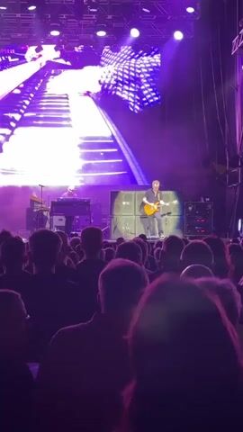 The video shows a concert performance by the band Deep Purple. A large crowd of people are silhouetted in the foreground, facing a stage illuminated with purple lighting. On stage, a guitarist is playing a solo on an electric guitar, with a large screen behind him displaying a visual of a keyboard. Other band members are visible behind the guitarist, including a keyboardist and a drummer. The overall atmosphere appears to be energetic and engaging.