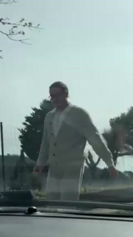 The video shows a person, presumably a man, with longish hair, a light beard, and glasses, wearing a light-colored cardigan, white shirt, and light-colored pants. He is standing near a white car and opens the driver's side door. The video is shot from inside the car. Outside the car, there are trees, a fence, and a clear sky. The man is not speaking in this section of the video.