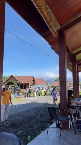 A vertical video begins with a view of a wooden covered outdoor seating area with two tables and chairs visible in the foreground. In the middle ground, a paved road runs horizontally across the frame, with several people and motorbikes on it. On the left side of the road, a group of people are gathered in front of a building with a traditional-looking roof. A man in an orange t-shirt stands on the left side of the road, facing the camera and giving a thumbs-up. On the right side of the road, a rice paddy field is visible. In the background, a large volcano looms under a bright blue sky with some clouds. The video progresses by panning slightly to the right, revealing more of the outdoor seating area and the people seated at the tables. The video ends with a view of the volcano behind the rice paddy field.