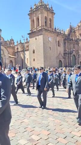 A large group of men wearing dark gray suits with blue ties are walking in formation across a cobblestone square. They walk toward the camera and away from a large, ornate stone church in the background, under a bright blue sky. Behind the men, a crowd of people is visible, along with a banner featuring a blue design. The church has multiple towers and a clock on its facade. The scene is bright and sunny.